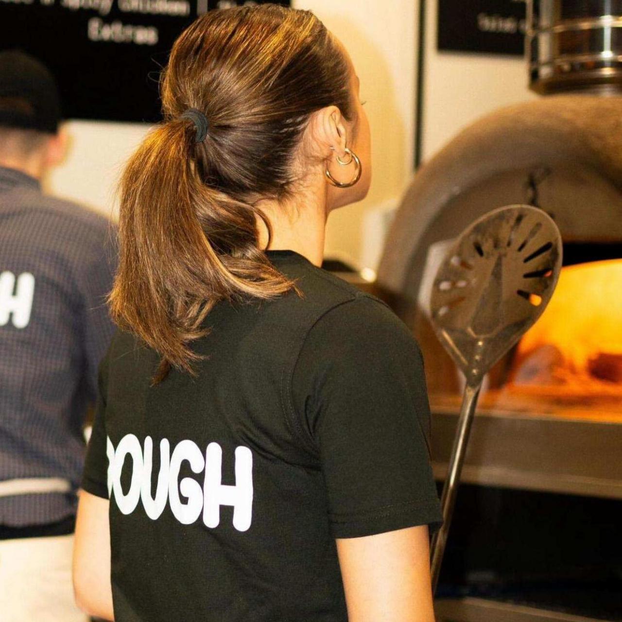 A young female wearing her hair in a ponytail and in a black T-shirt with 'DOUGH' written in white capital letters on the back. She's stood facing a pizza oven in the store.