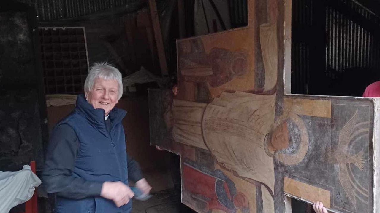 Vicar smiling at the camera while the painting is carried on its side out of the barn 