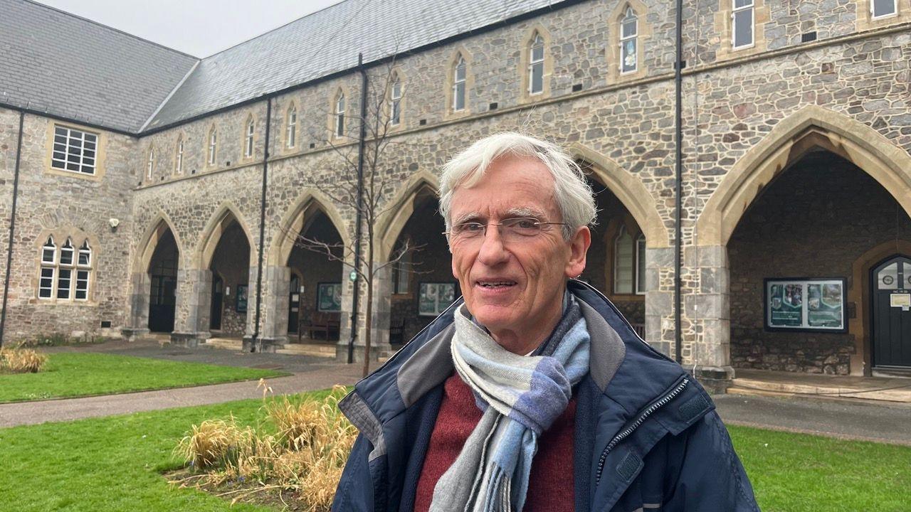 Brahm Norwich is Emeritus Professor of Education at the University of Exeter. He is standing in front of a university building and is wearing a blue coat and a blue scarf with a scarlet-coloured jumper.