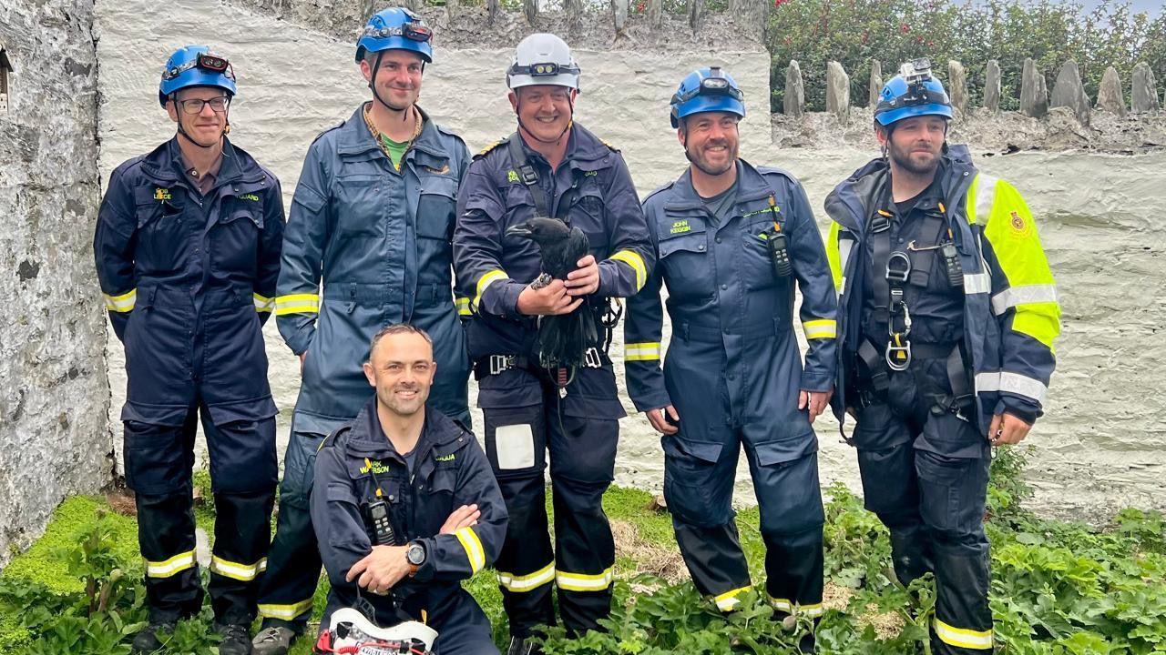 Six coastguards all stand together and smile, one holds the baby raven.