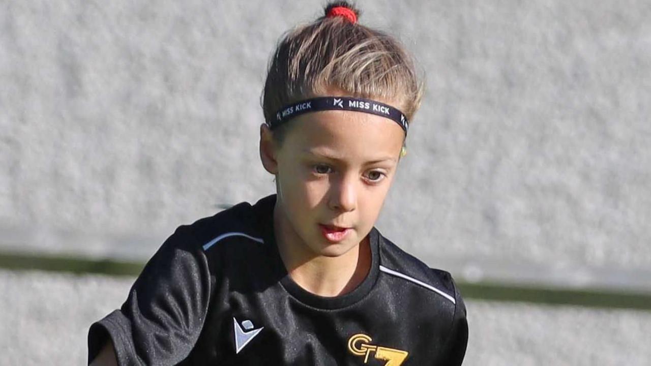 Poppy playing football. She has dark hair pulled back into a ponytail and is wearing a black football kit. She is looking down at the ground