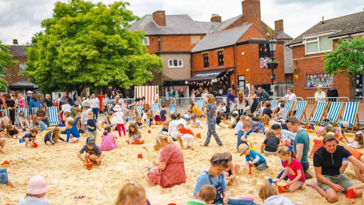 A large number of children and adults sat on a large, rectangular sandy area in a town, with blue striped deck chairs surrounding the sand and red brick buildings in the background