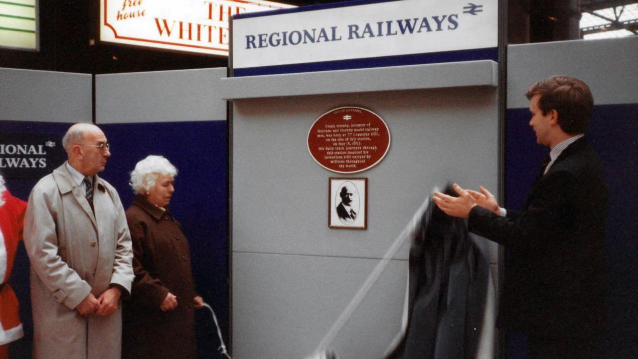 A dated photo of the original plaque as it was unveiled 30 years ago. A man can be seeing pulling a way a black curtain, revealing the sign, while two older people watch on.