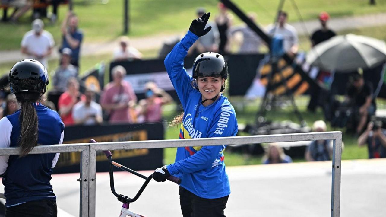 Queensaray with dark hair wearing a black helmet and blue top and holding a BMX bike