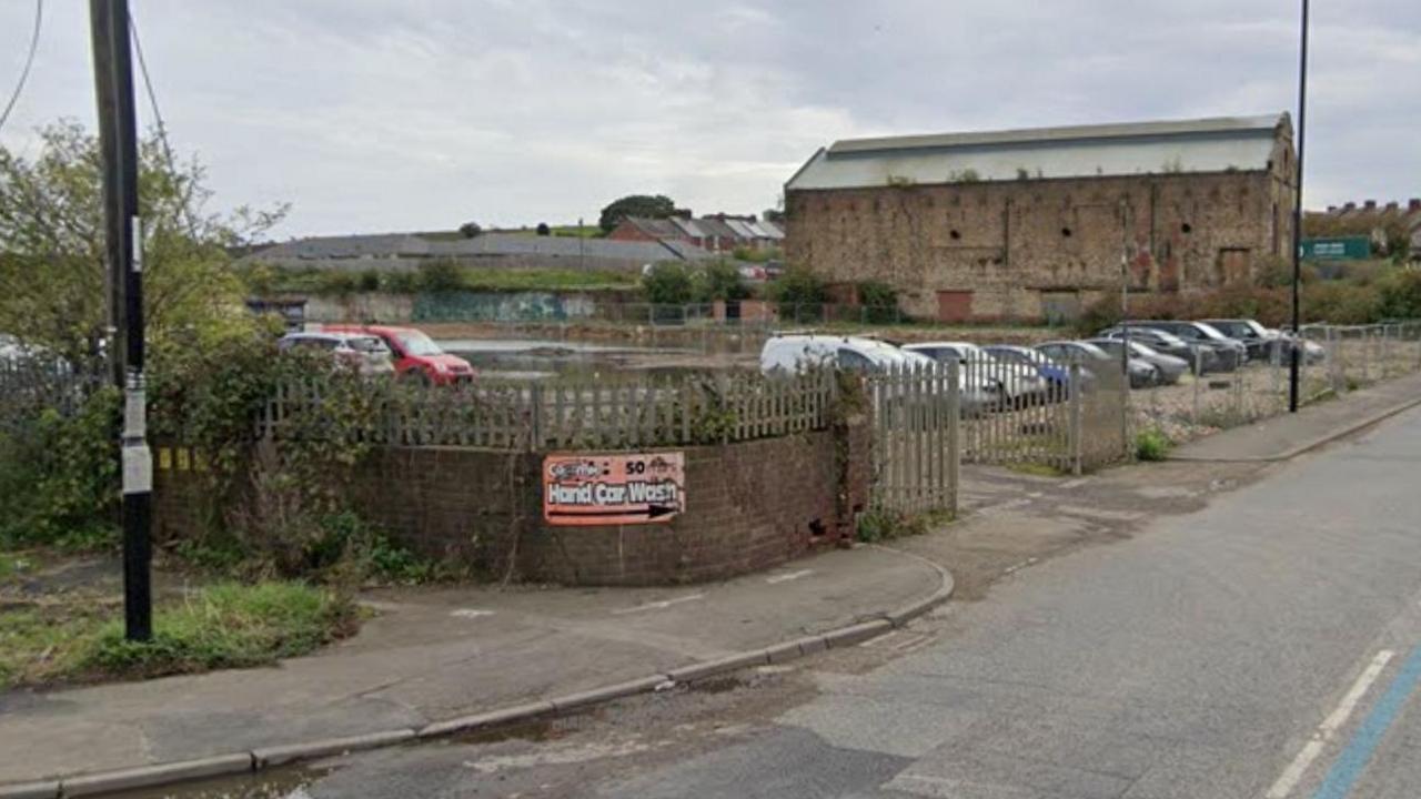 A derelict area of land off Philadelphia Lane. A wall and metal fencing surrounds the area which is being used as a car park. Behind it stands a large, barn-like brick building with pointed roof.