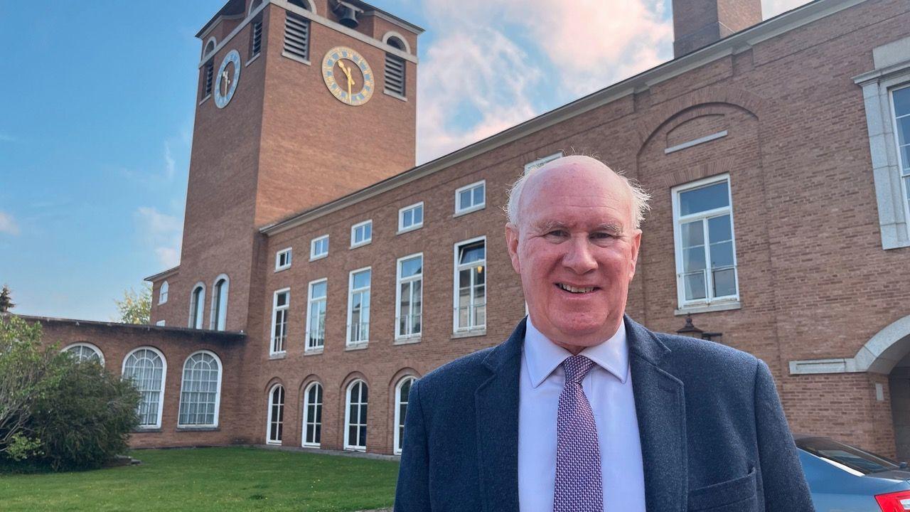 James McInnes, leader of the Conservative-run Devon County Council, in a suit outside County Hall. He is smiling directly at the camera.