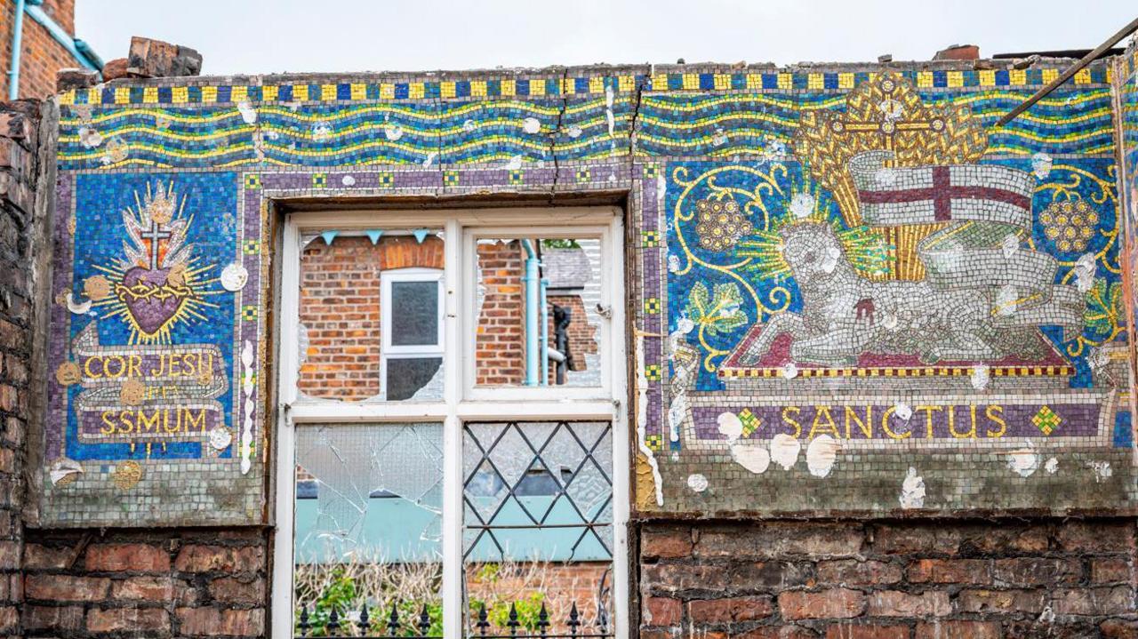 A broad, horizontal mosaic straddles a broken window on a partially demolished wall. The left side of the mosaic shows the Sacred Heart of Jesus, while the other shows a lamb with the flag of St George waving above.