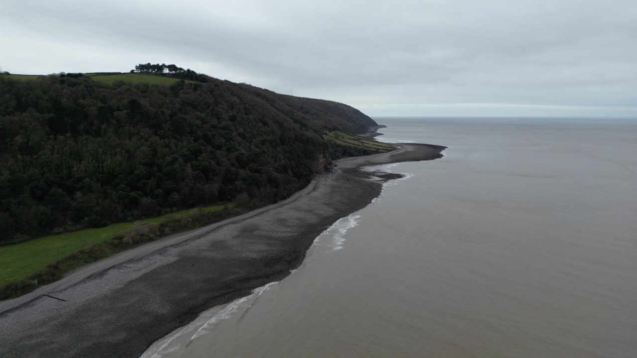 The coastline by Exmoor National Park seen from above. The sea is grey and there is a forest above the beach.