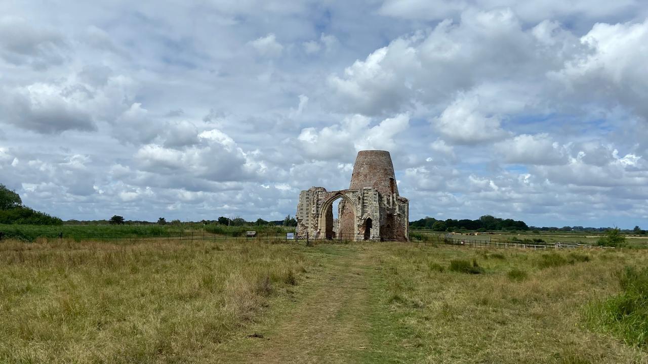 The ruins of a monastery in a large field. There is a stone gateway with partially collapsed walls. Behind it is a large round red-brick building.