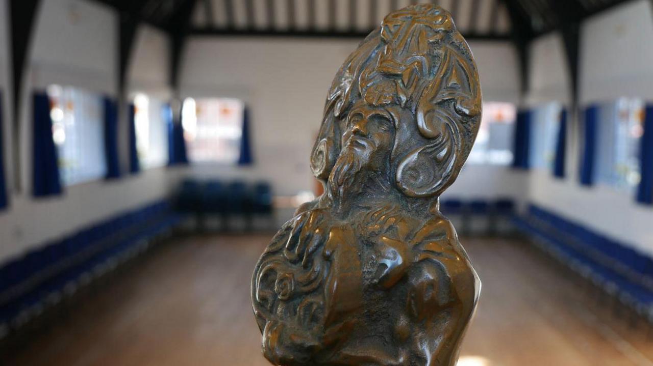 A closeup of the head and shoulders of the Reih Bleeaney Vanannan trophy, which is a statue of the sea god Manannan. It is in a white room with multiple windows that have navy blue curtains either side and wooden floorboards. 