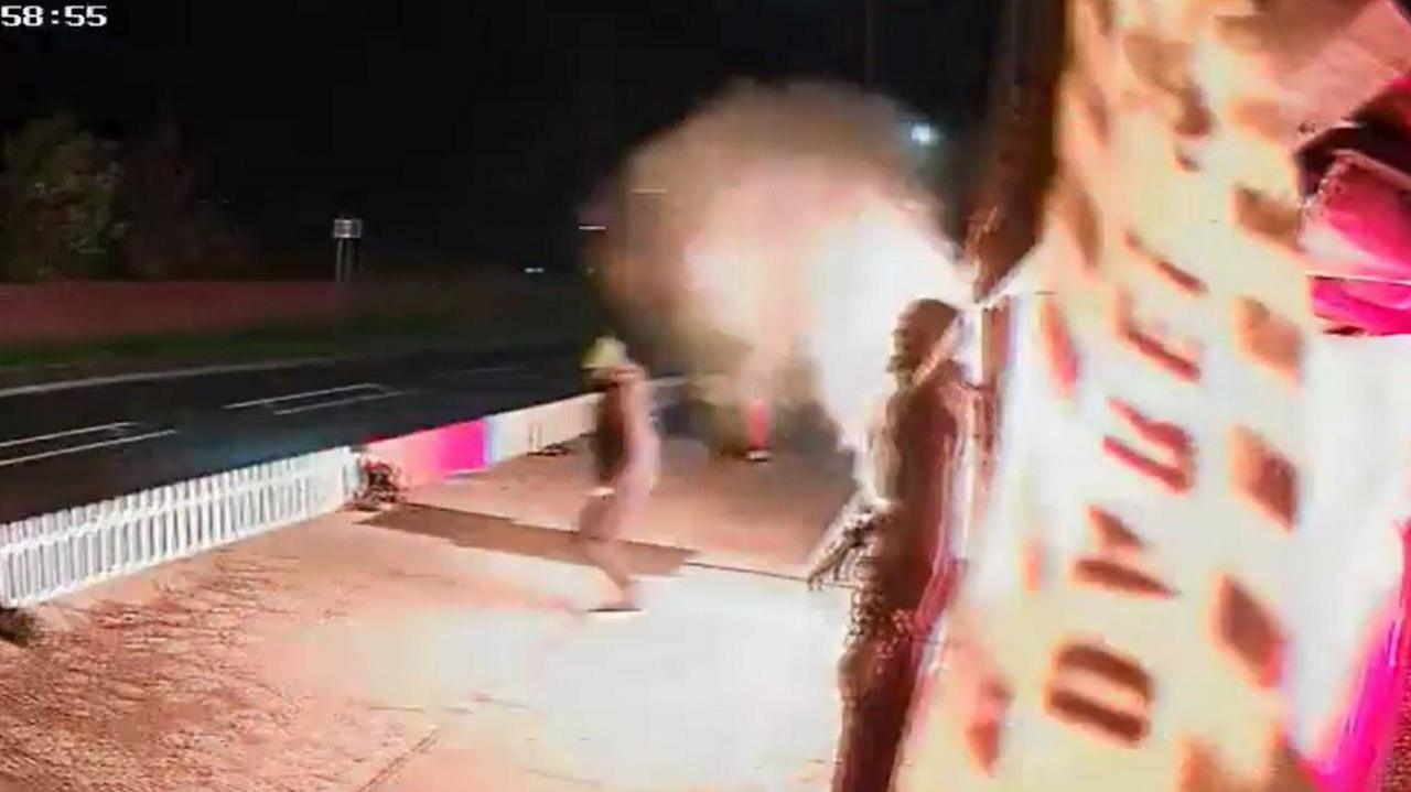 CCTV footage of the moment the display caught fire. A man in a hat and black coat is moving away from the display. An explosion of flames has erupted behind him from the display. A statue of a monster can be seen in the foreground.