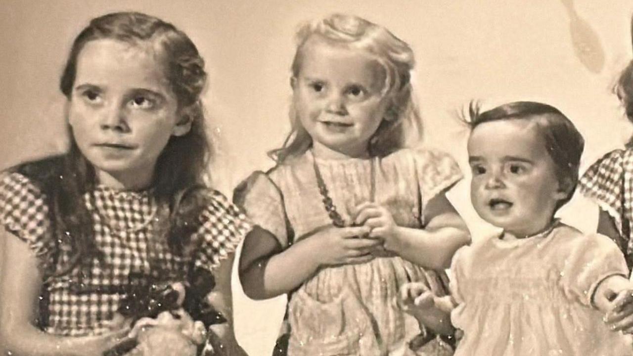 Old yellowing black and white photo of the three sisters as young children. The eldest on the left is wearing a checked dress and has long brown hair. The middle girl is younger and has blond hair and is wearing a plain dress while holding a necklace around her neck. The youngest on the right is a toddler with short dark hair.