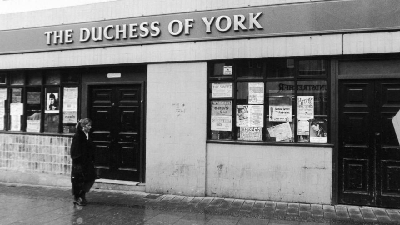 The front of a building with posters on the windows and a sign above saying The Duchess of York. There is a woman wearing a long black jacket walking past.