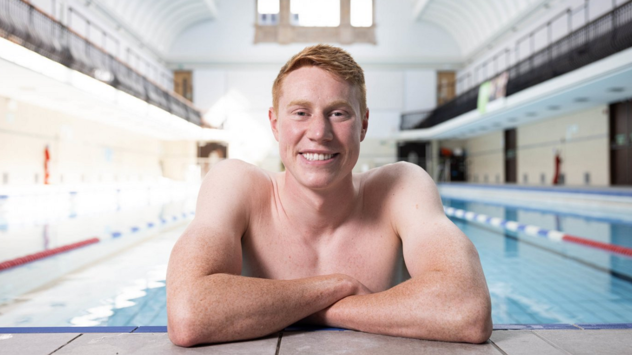Tom Dean in a swimming pool in a close-up shot of him leaning against the side of the pool, with arms crossed, and smiling to camera