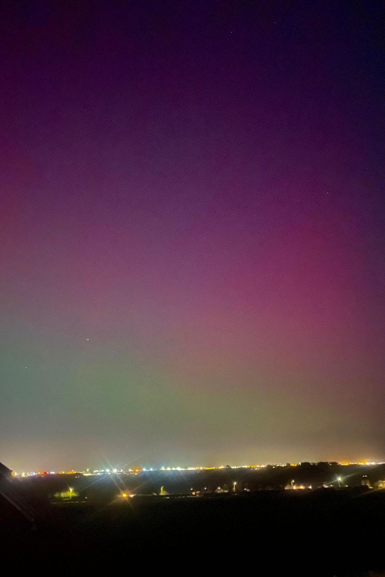 A shot taken into the distance, with the green and purple of the Northern Lights seen above lights from houses on the horizon.