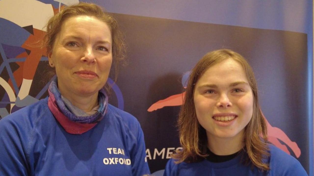 Sarnia and Blossom Martin both smiling and have medium length brown hair. They are stood in front of a purple board.