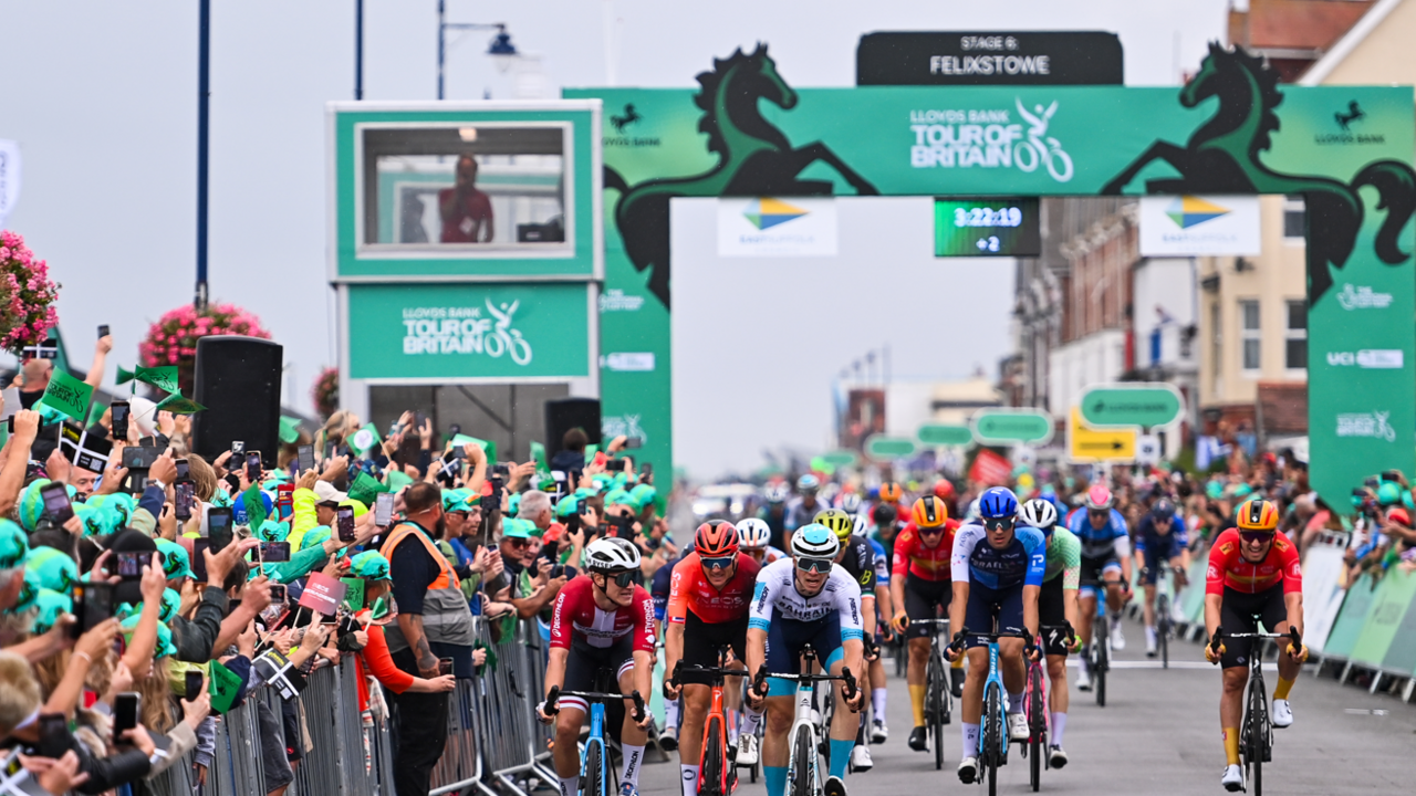 Cyclists are coming towards the camera on a road. There are metal fences either side of the road where people are standing watching. A sign goes over the road which is green and has the horse logo for Lloyds.