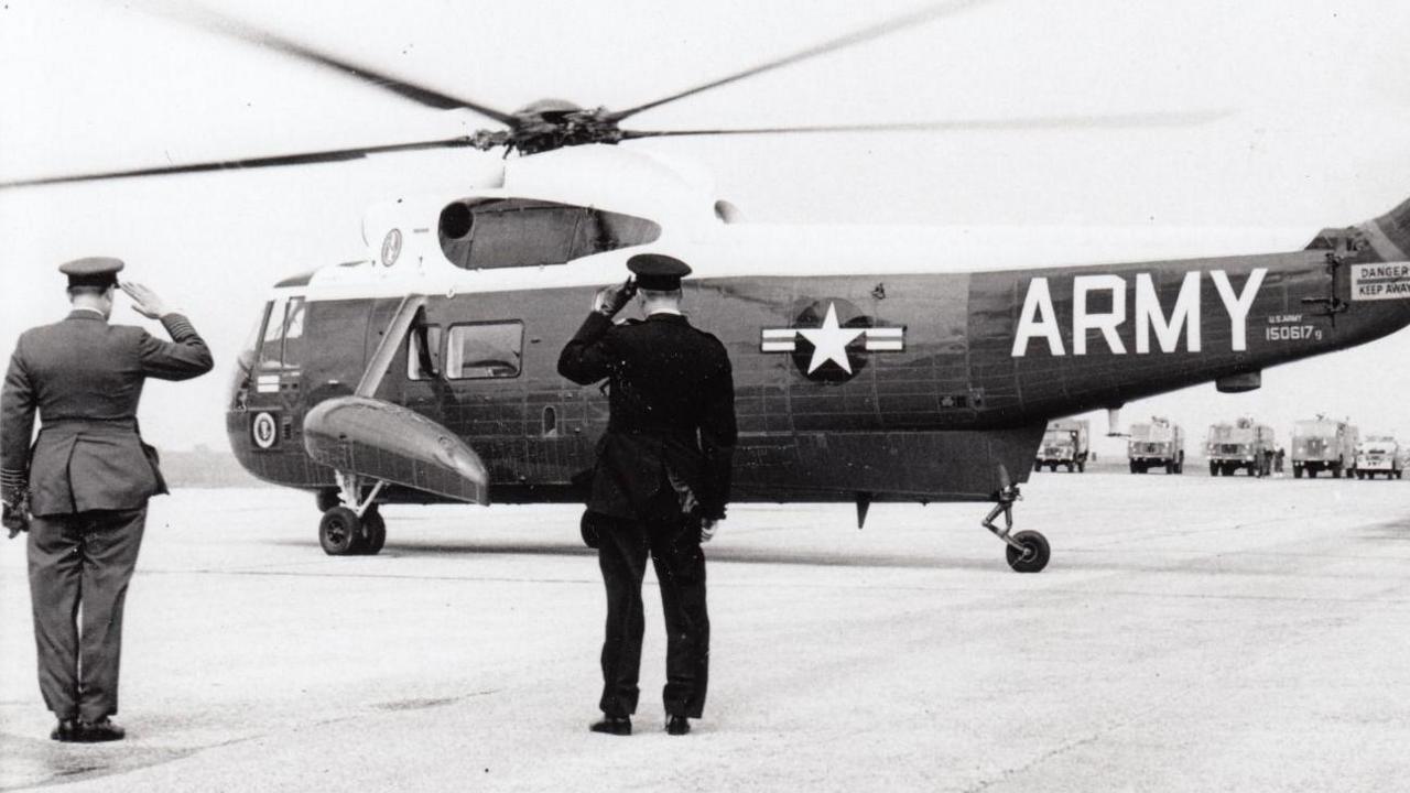 A black and white photograph of a US Sikorsky military helicopter about to take off. Two men stand nearby, both are saluting.
