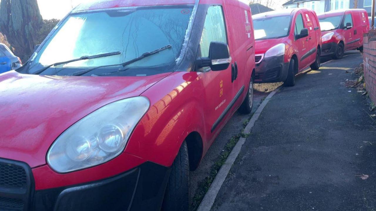 Three Royal Mail vans are parked bumper-to-bumper on a residential street. Two are parked on the kerb. Terraced houses are in the background.