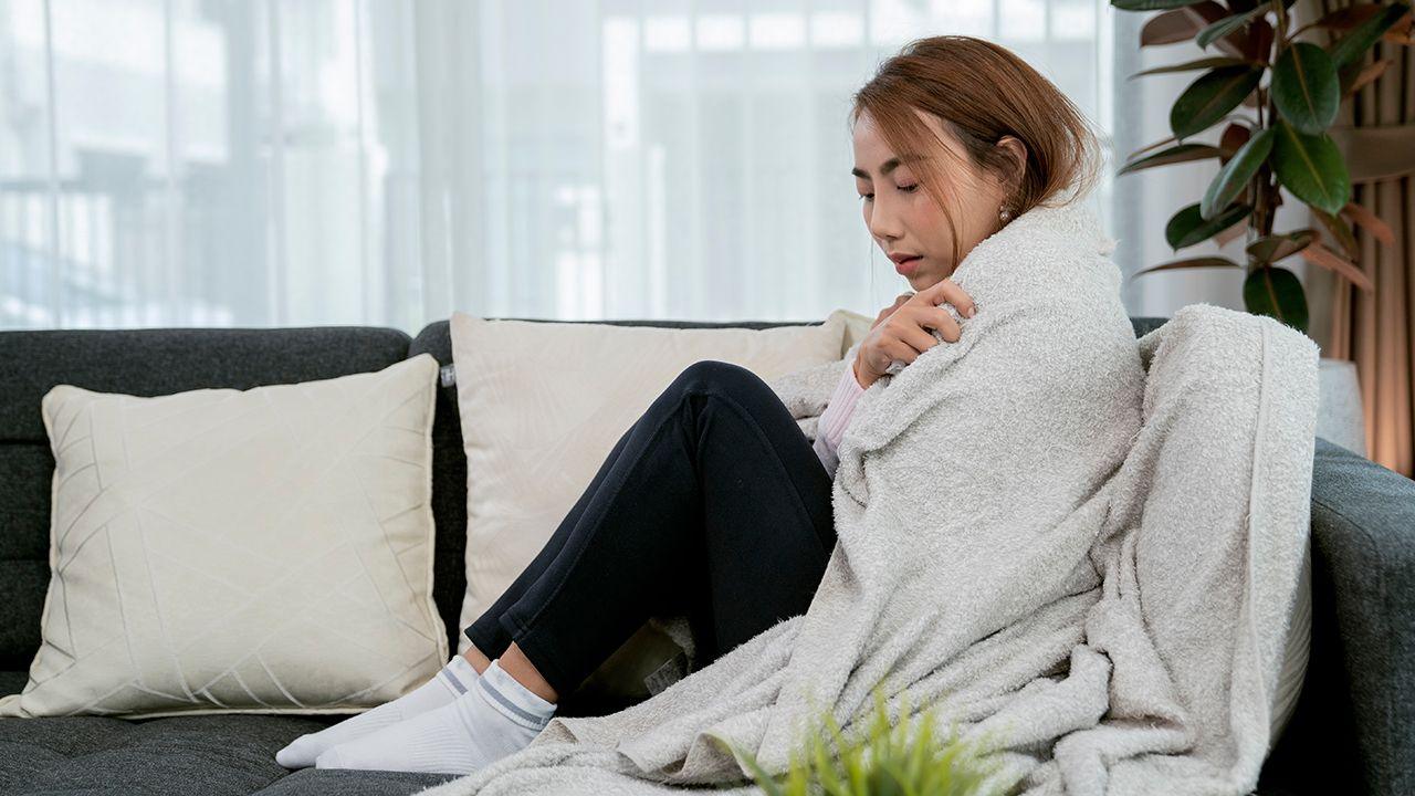 A woman sits on the sofa with a blanket wrapped around her body to keep warm.