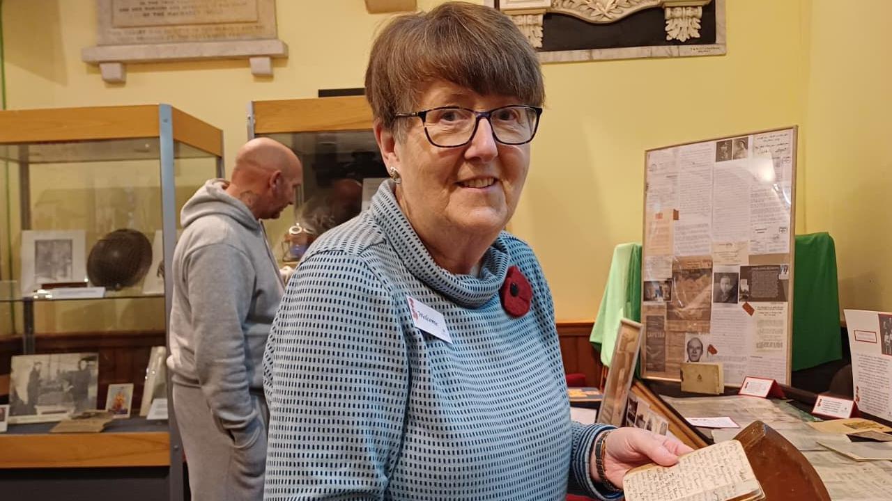 Rosemary is holding a diary. She's wearing a knitted poppy. She wears glasses.