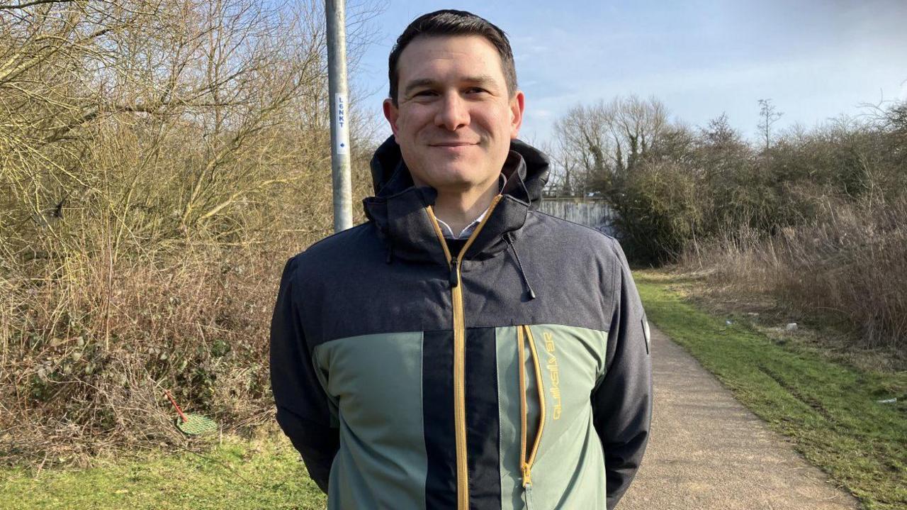 Smiling man wearing a black and green weatherproof jacket. He is standing on a footpath. Behind him are bushes and trees.