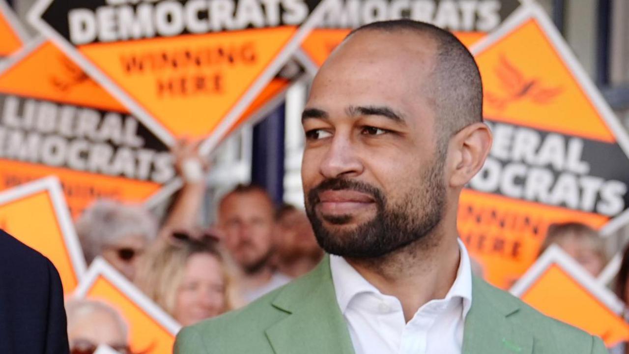 Josh Babarinde MP campaigning in Eastbourne at the general election in July 2024. Behind him are a series of Liberal Democrat campaign signs.