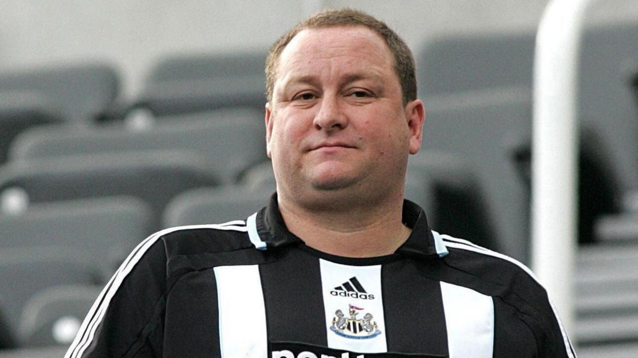 Mike Ashley is wearing a Newcastle United shirt. He has short brown hair and is clean shaven.