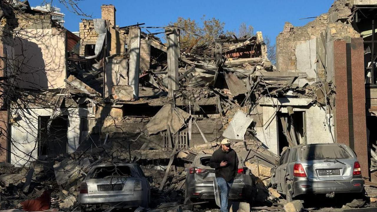 Destruction from missile attack on Kharkiv - man walks towards camera in front of three cars and a wrecked building