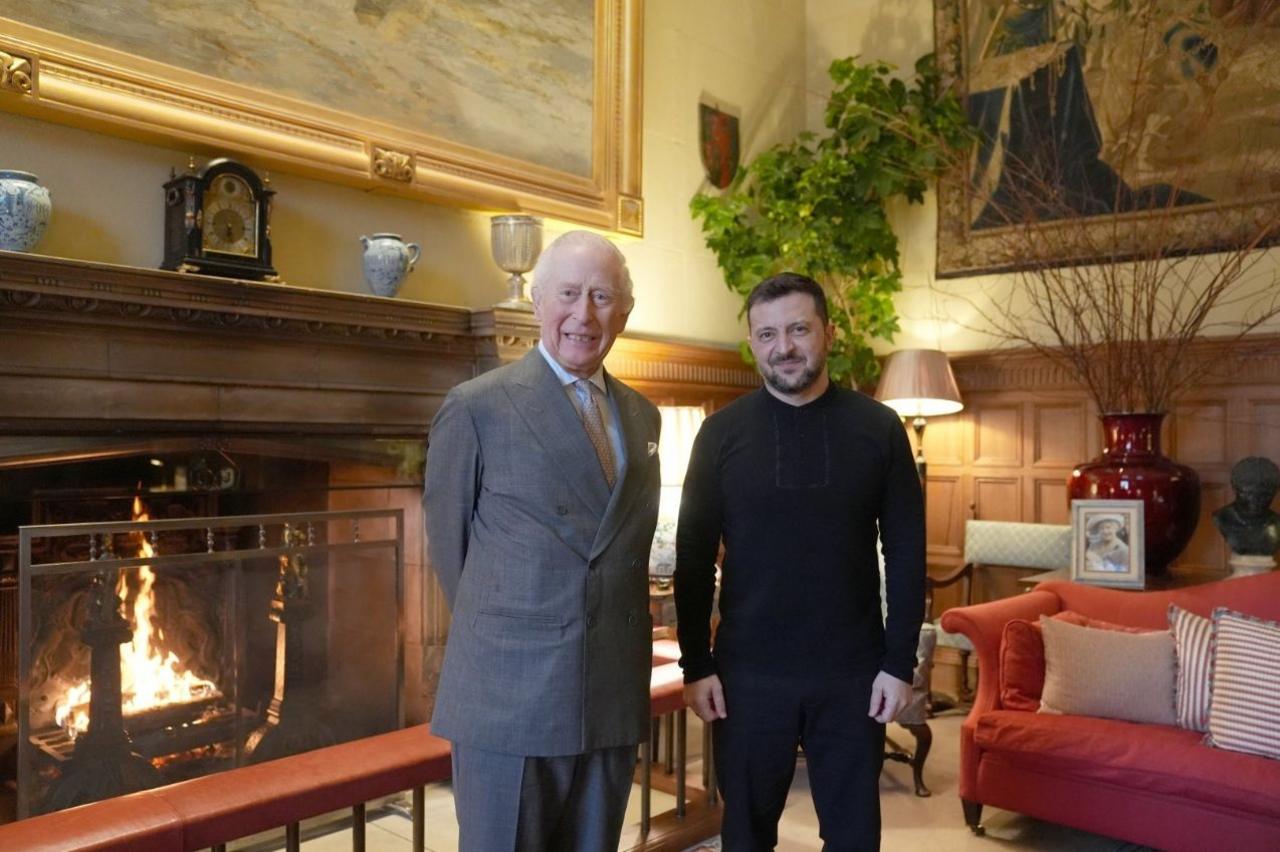 King Charles wearing a grey suit standing next to Volodymyr Zelensky in a wood-panelled sitting room with a fireplace and red sofa with striped cushions.