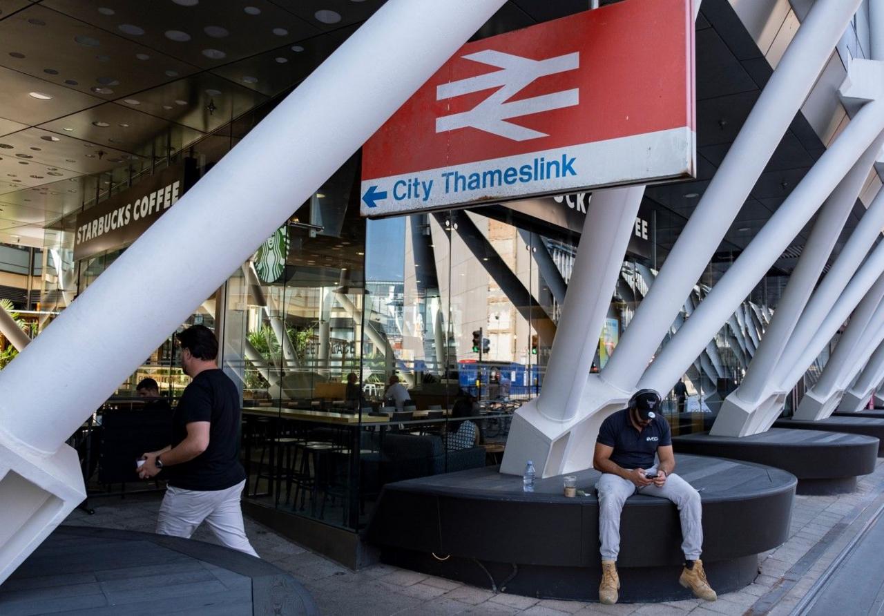 A railway station with a large sign bearing the railway symbol and the words City Thameslink. There are white roof support poles in the foreground, and a Starbucks Coffee in the background. A man is sitting below the sign looking at his mobile phone.