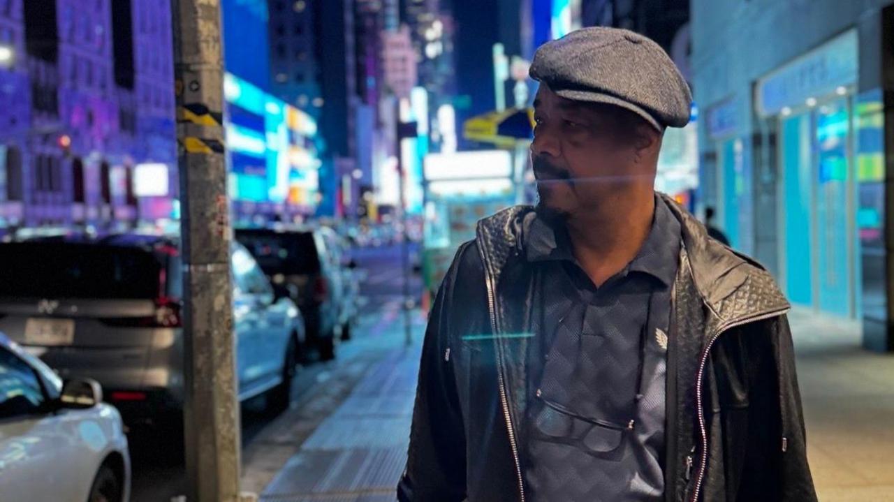 Earl Falconer, wearing a flat cap and leather jacket, walks on a street in New York City at night with buildings behind him, bathed in blue light, and a series of parked cars on his right.