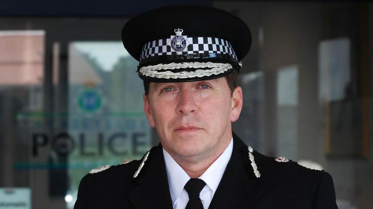 Chief Constable Carden has short dark hair and blue eyes and is wearing a police uniform and looking into the camera. He is standing outside Merseyside Police building.