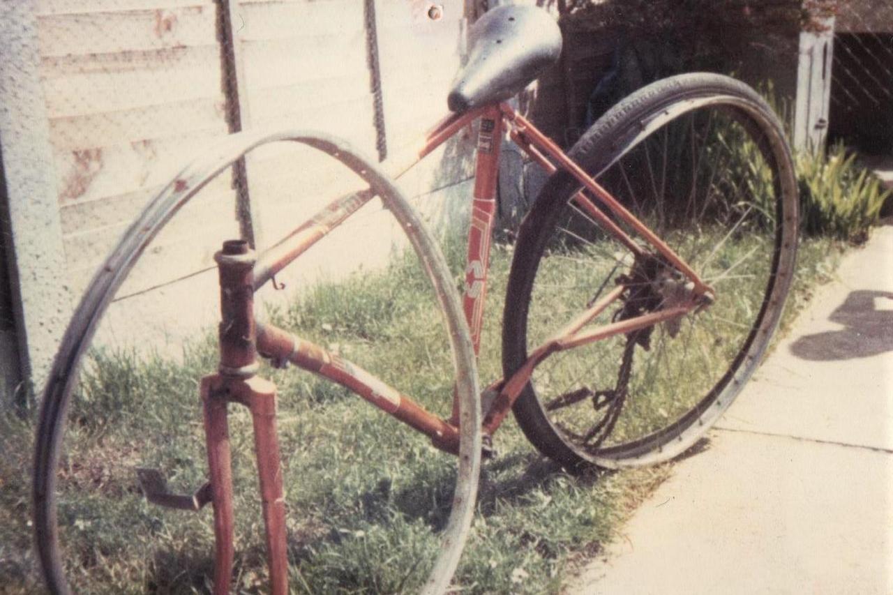 An old photo from 1982 of Mark Williams' damaged bicycle. It is a red road bike with a slightly damaged back wheel and the chain broken. The front wheel is missing, and the metal wheel rim with no spokes is leaning up against the frame of the bike. 