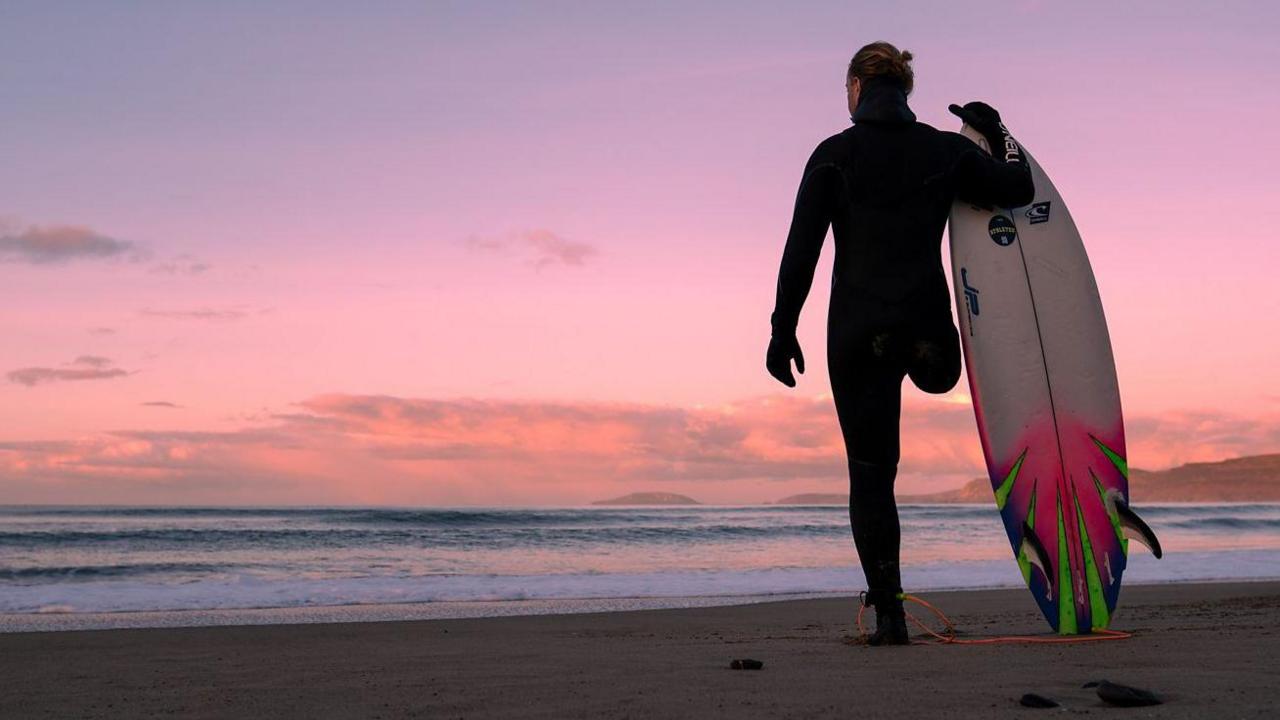 Surfer in a full body black wetsuit and gloves. He has one leg and he stands on a beach at colourful cool pink sunset. He has his back to the camera with the sea in front and the vast sky beyond. He's holding a white and shocking pink and green surfboard with its tip pointing skyward and the other end resting on a sandy beach.