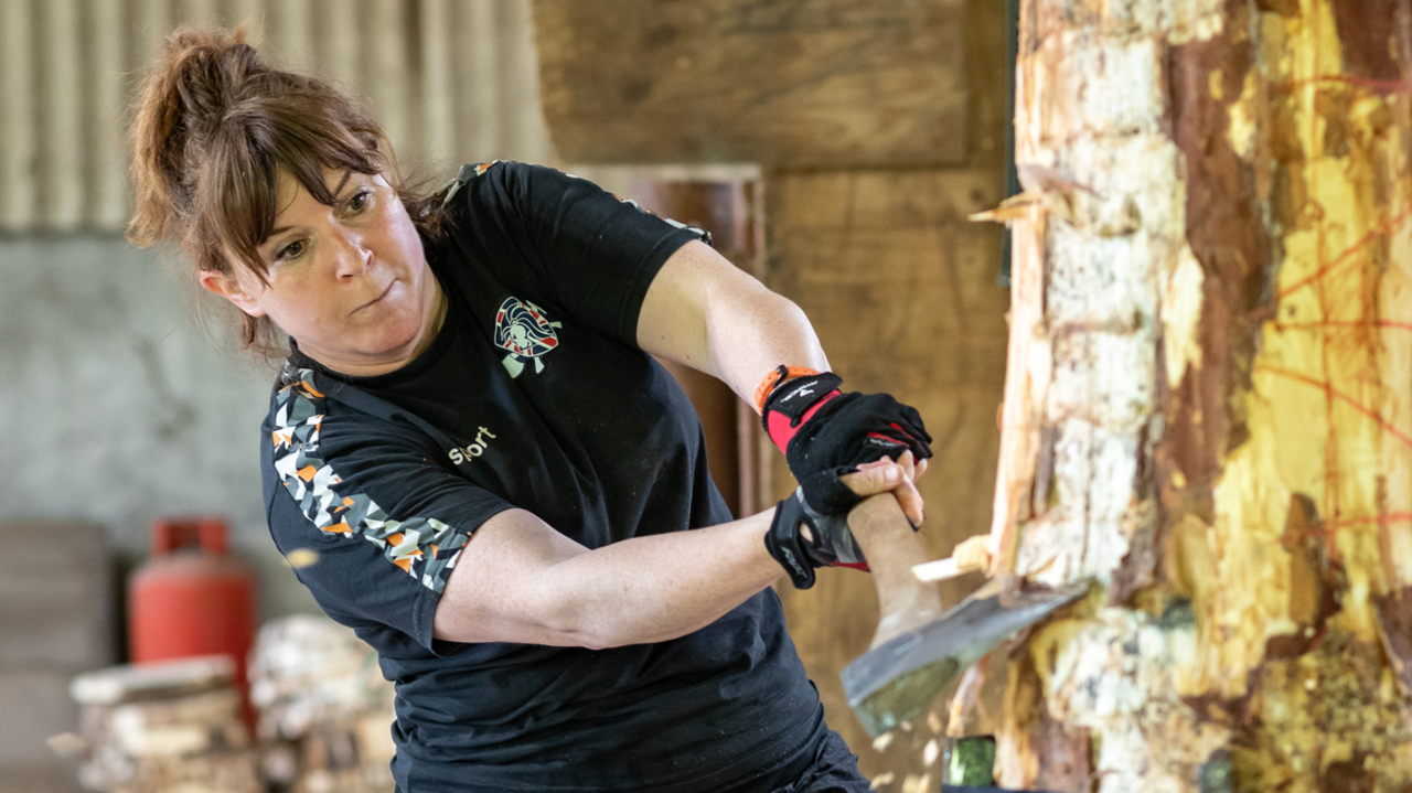 A woman with brown hair that has been tied back wears a black t-shirt while she uses an axe to chop into a large log.