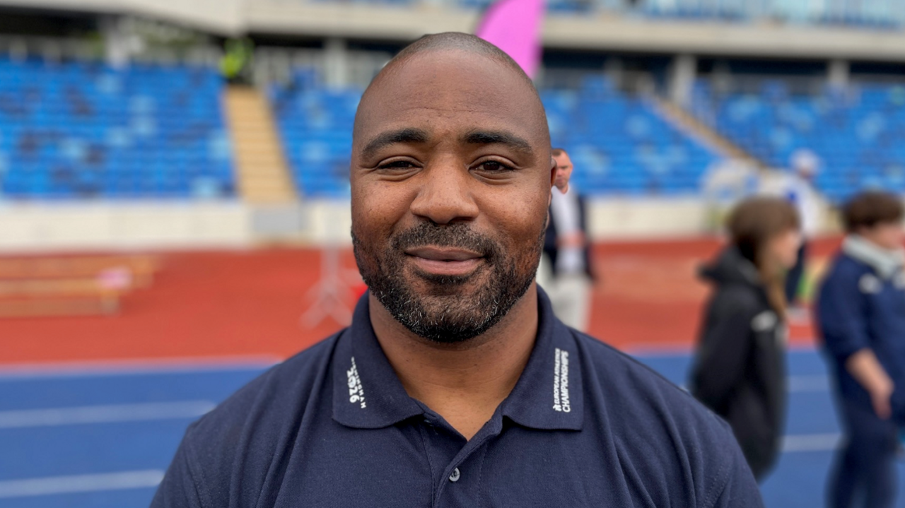 A headshot of Mark Lewis-Francis wearing a blue polo shirt stood in front of a blue race track. He has shaved dark hair on his head and a short, black beard.