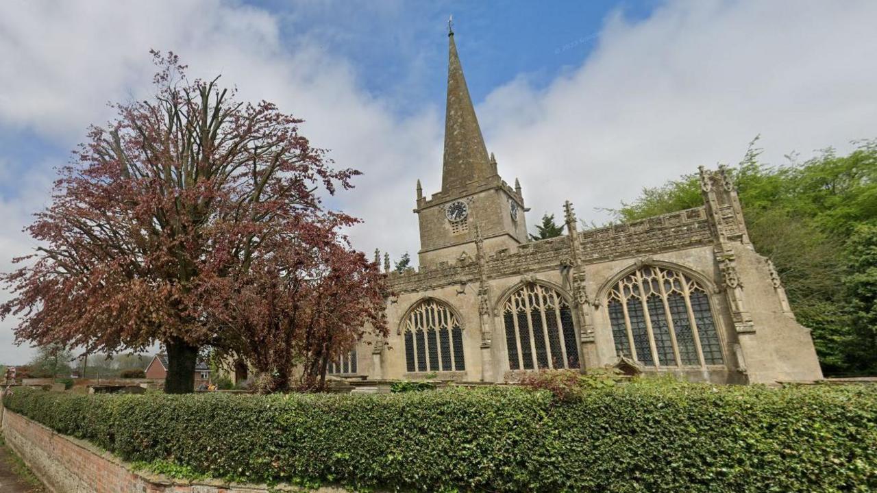 A view of St Nicholas's church in Bromham, where many of the Bayntun family are buried. Sunny day with a green hedge outside and large tree with reddish leaves to the left