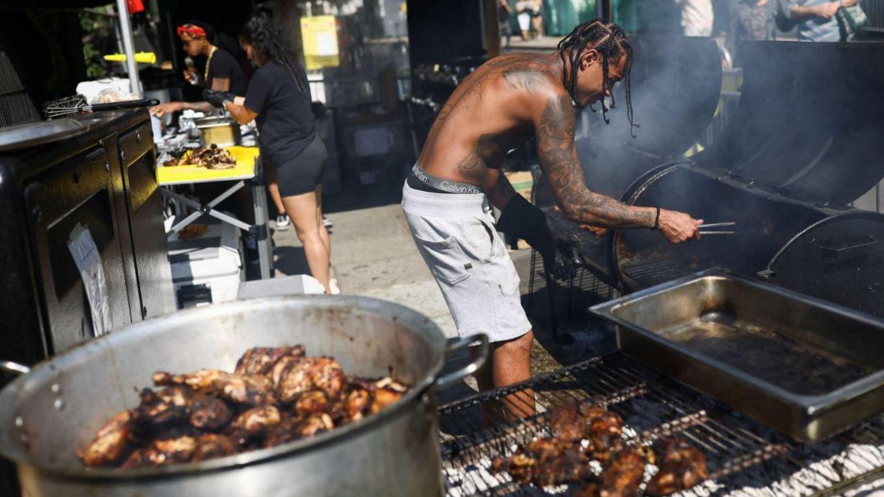 A man is bent over a barbecue with smoke coming out of it. In the foreground is grill with a meal dish of barbecued meat inside.