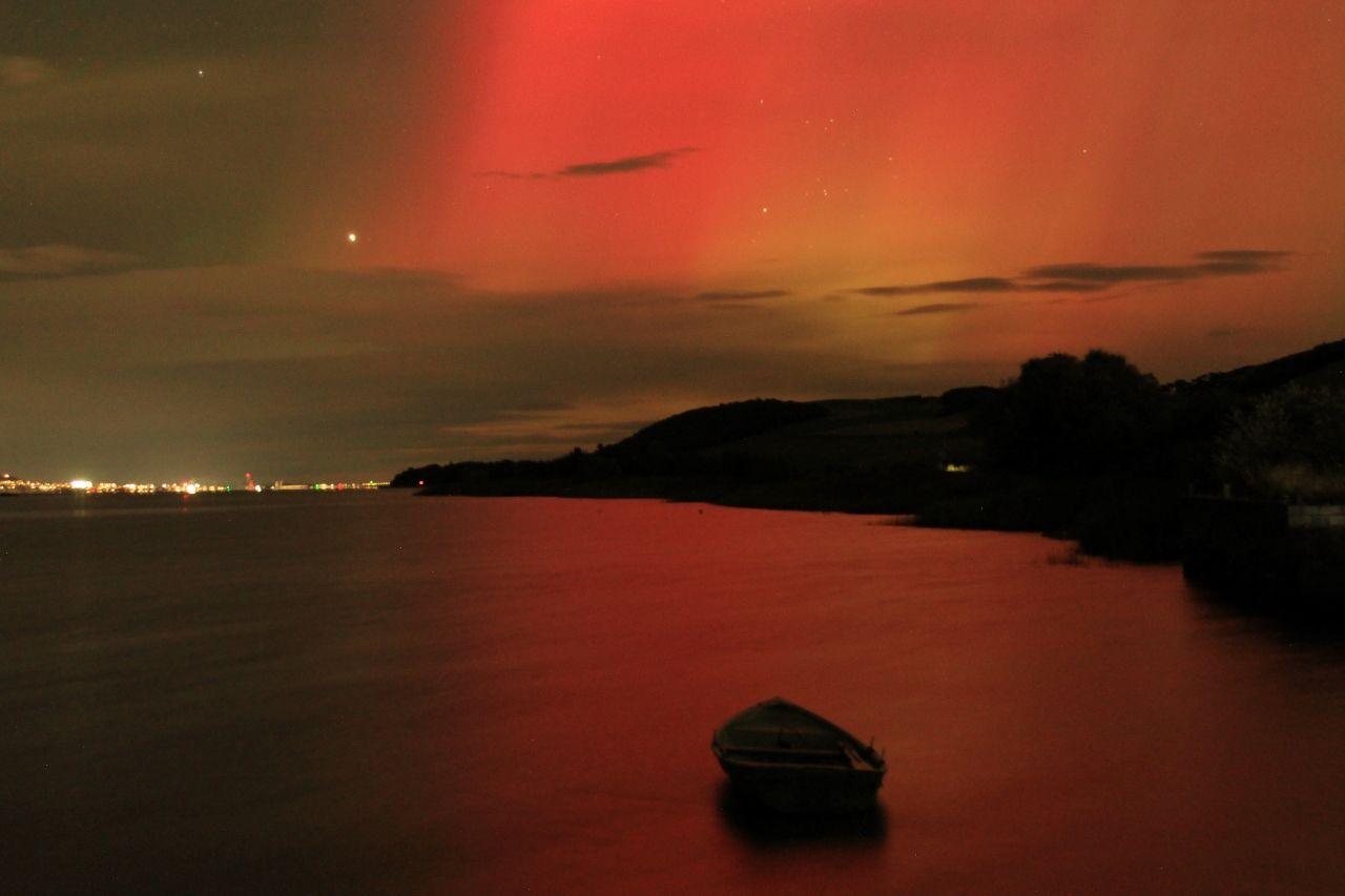 A red aurora sky illuminates the River Tay. A small boat sits in the water in the foreground. The lights of Dundee are visible in the background.