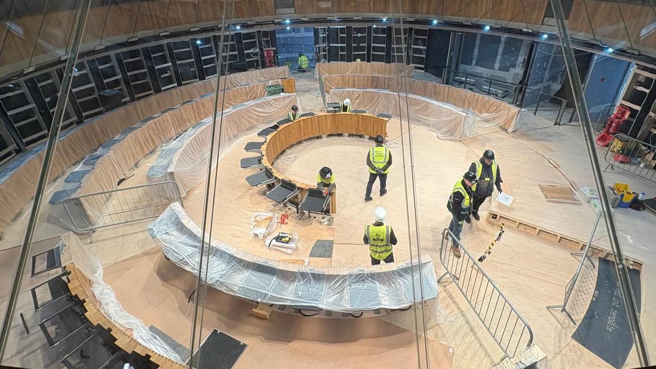 Llun o weithwyr yn brysur yn gweithio yn siambr y Senedd. Bydd nifer y desgau a'r offer technegol yn cynyddu i ddarparu lle i'r Aelodau Senedd newydd fis Mai.