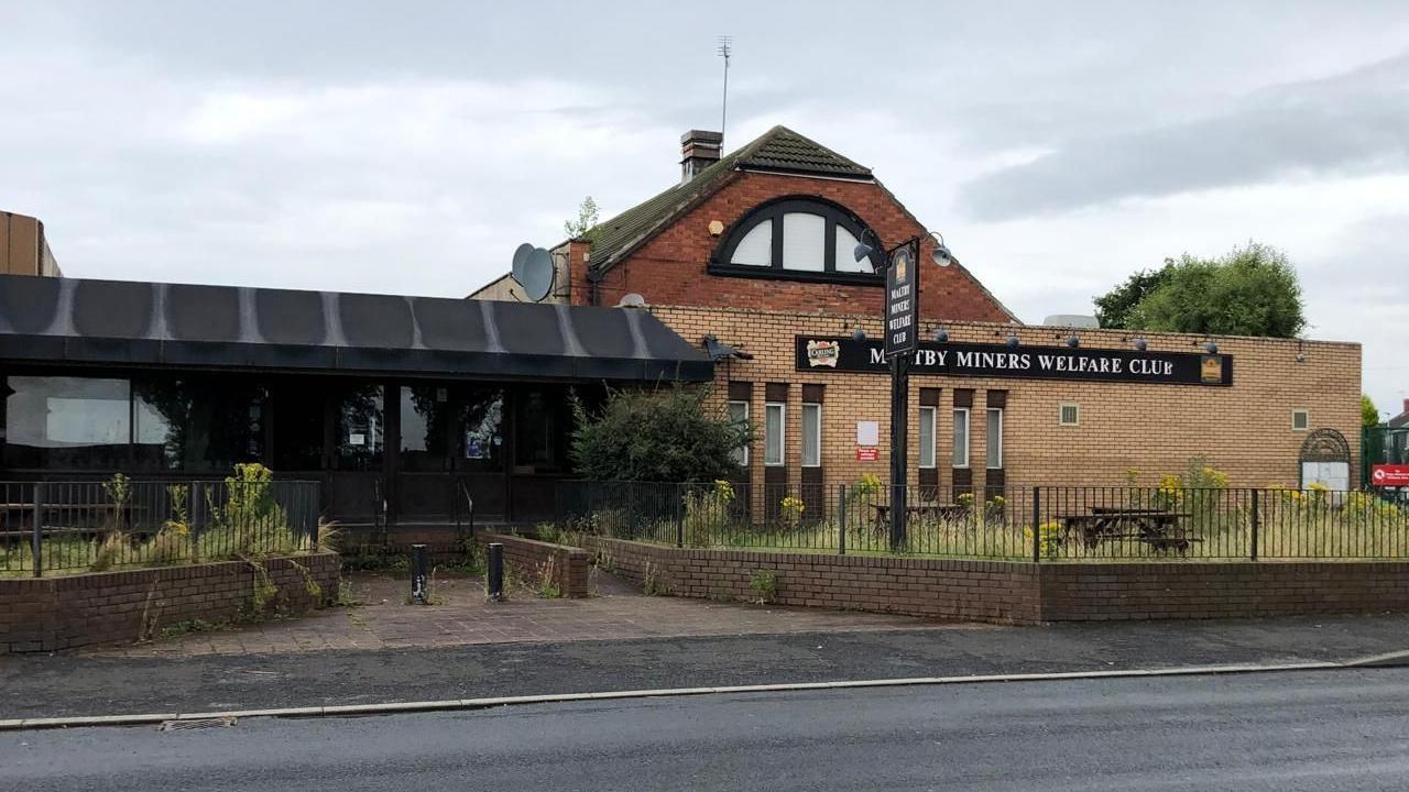 Maltby Miners Welfare Institute was a brown and red-brick building with a large black and white sign across the entrance. A patch of long grass and wooden picnic benches can be see outside.