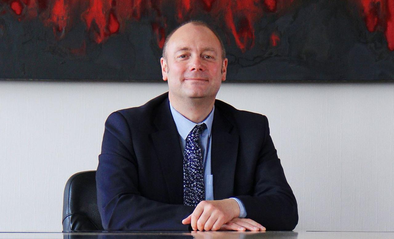 Darren sits at a desk with his hands folded on top of one another. He smiles at the camera and is wearing a dark blue suit with a floral navy tie.