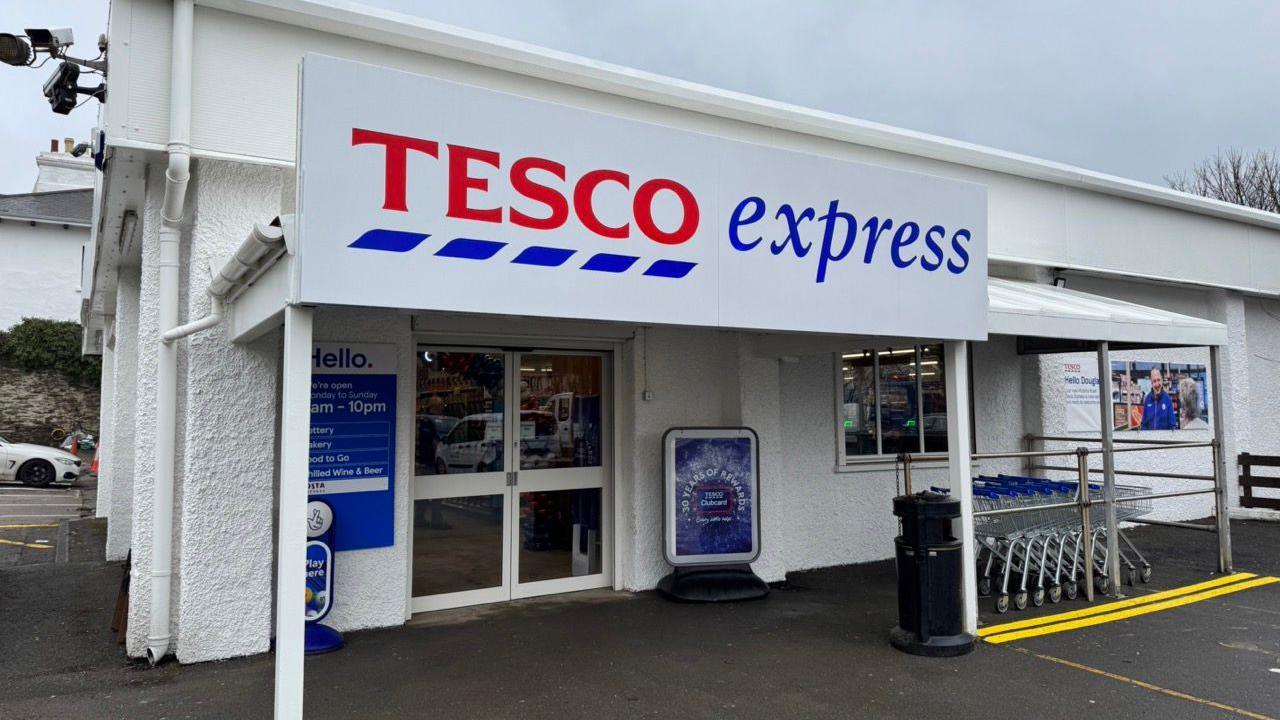 The exterior of the new store, which is a white building with a sign that reads Tesco express in red and blue on a white background. A row of trolleys are lined up on the right of the large glass double doors at the entrance.