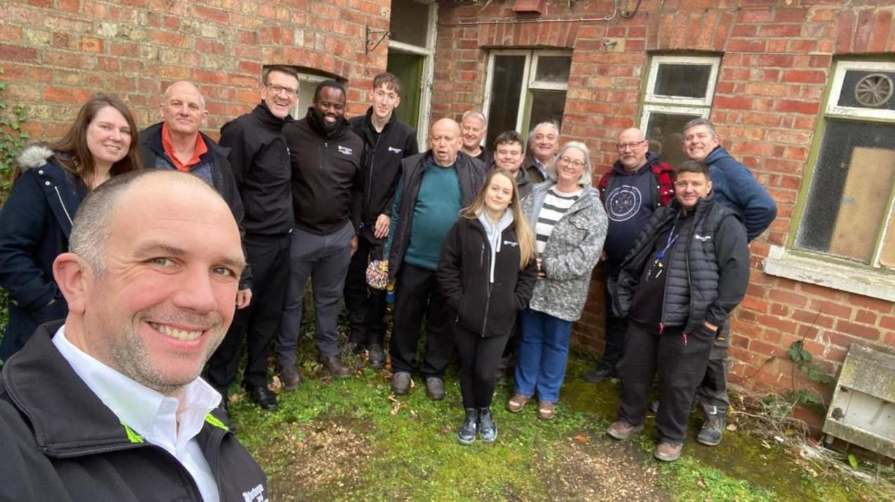 15 people are looking at the camera as the man in front takes a selfie. They are dressed in winter clothes, coasts and boots. They are all smiling and are stood outside the abandoned cottage.