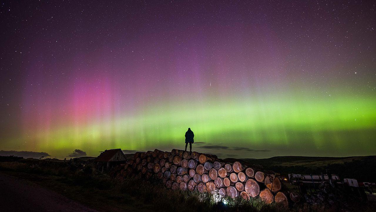 A person stands on a pile of logs in a rural part of the Highlands, they are silhouetted against the night sky with the purple and green of the northern lights in full view amongst a few stars