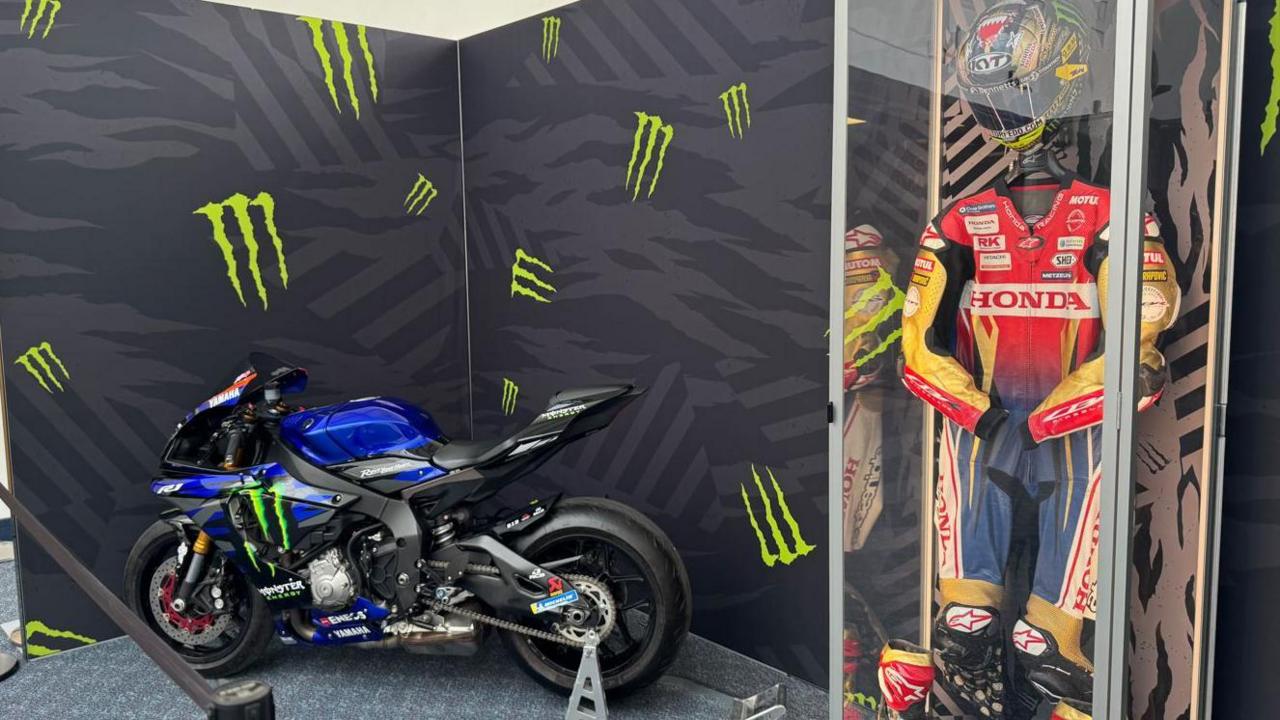 There is a blue motorbike on the left and a st of red, blue and gold leathers in a display case on the right with a helmet.