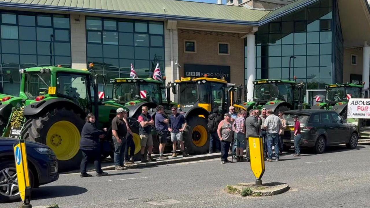 Farmers stand near tractors parked on a pavement outside a glass-fronted building