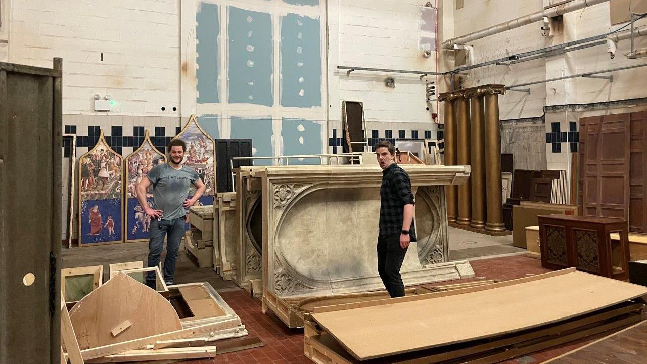 Two film students standing in a large room surrounded by pieces from the set of the drama Wolf Hall. It includes a wooden chair, some gold pillars and arches lying on their sides.