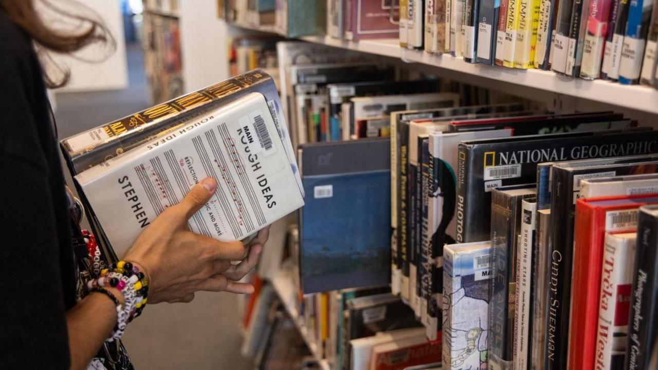 A woman's hands holding on to two books, in front of shelves holding lots more books